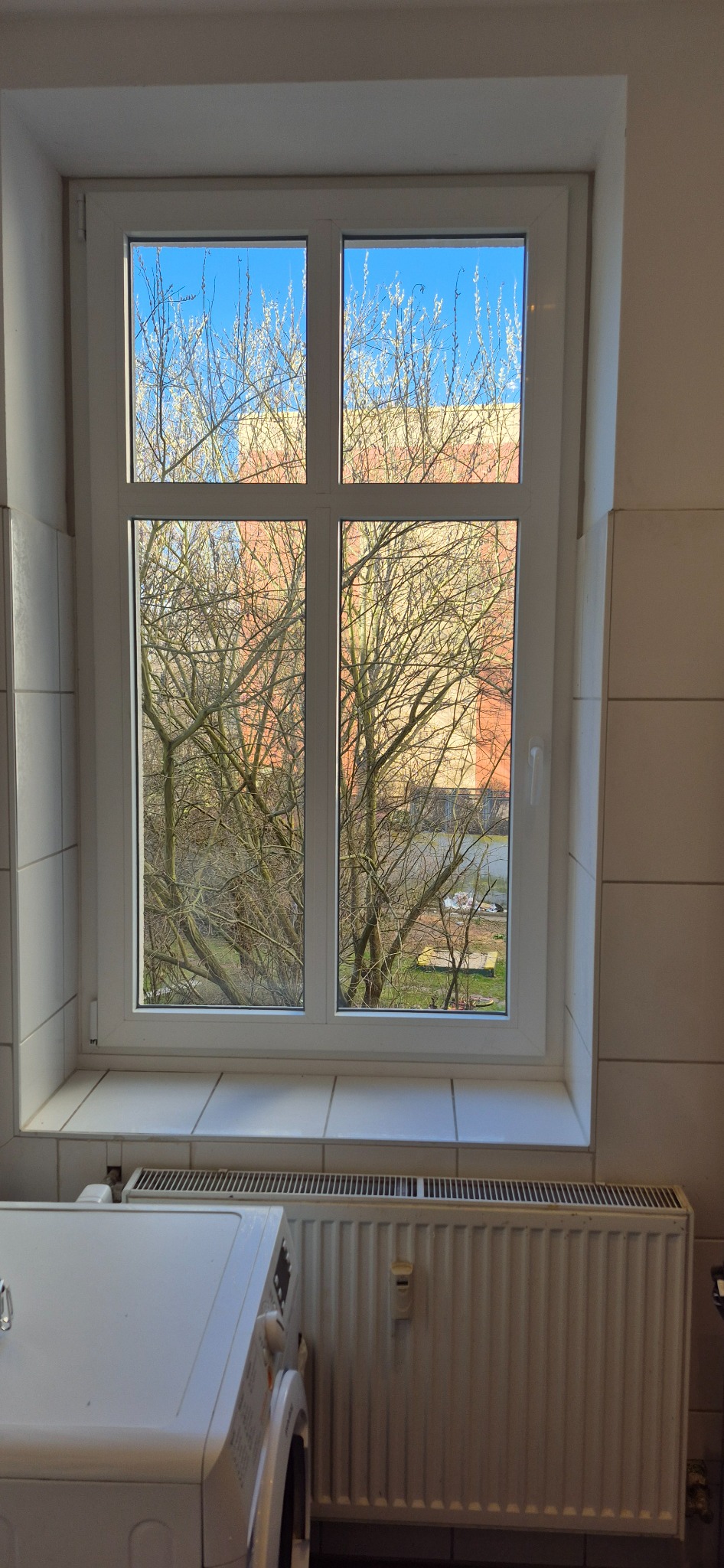 Białe okno PCV z widokiem na drzewa i budynek. W tle grzejnik i pralka, wnętrze wykończone płytkami. Widok z okna na błękitne niebo.