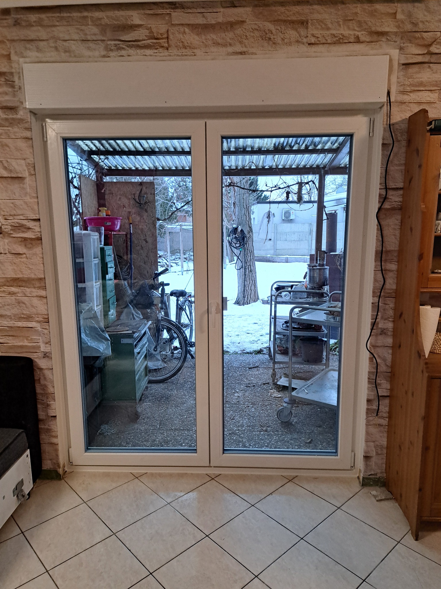 Białe, dwuskrzydłowe drzwi balkonowe z roletą zewnętrzną, widok na zadaszony taras z rowerem i wózkiem narzędziowym w zimowej scenerii.