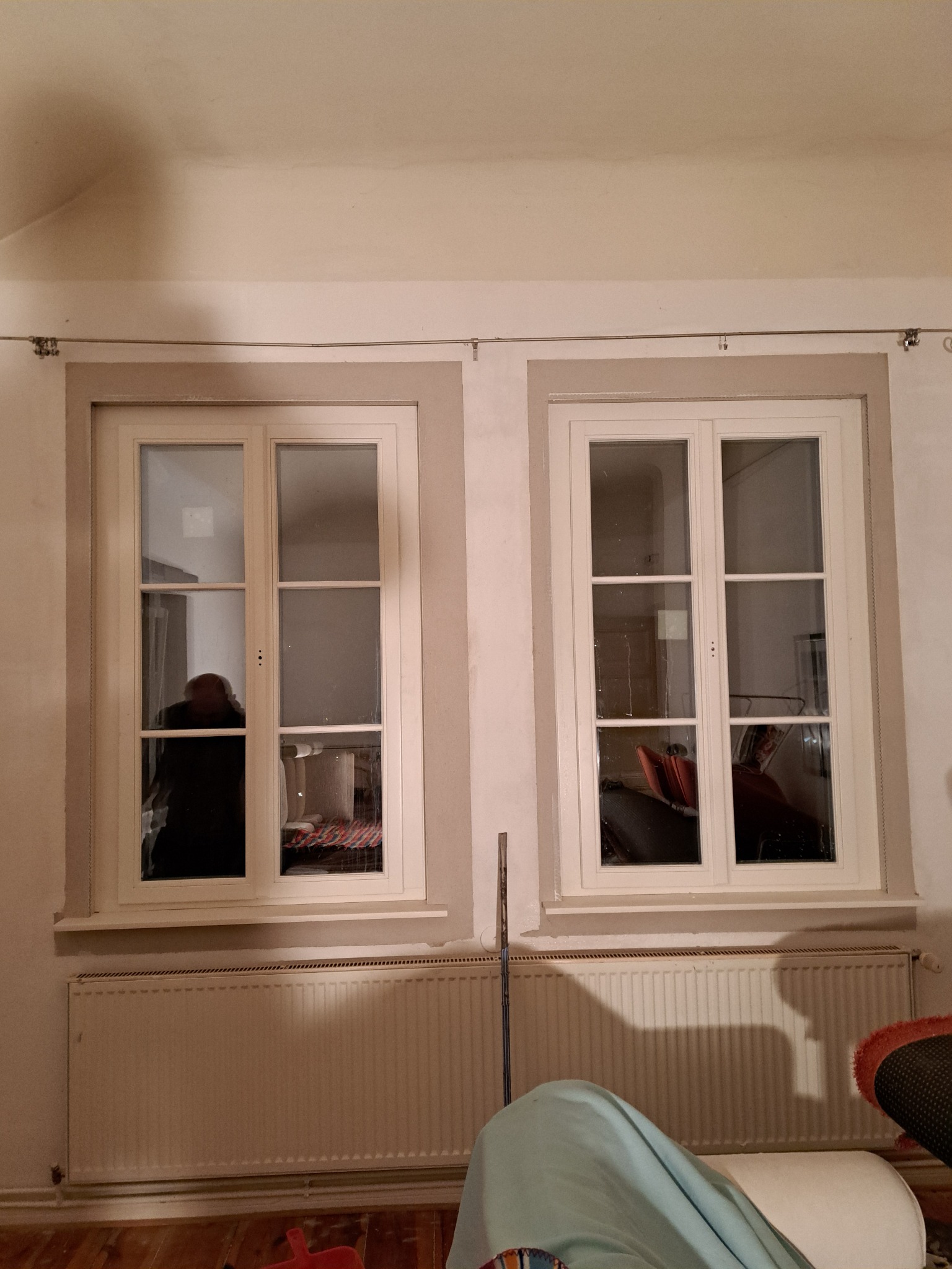 Dwa białe okna z szprosami w pokoju, widoczny grzejnik i odbicia w szybach. Okna osadzone w jasnych, beżowych ramach. W tle widoczne meble i inne elementy wyposażenia.