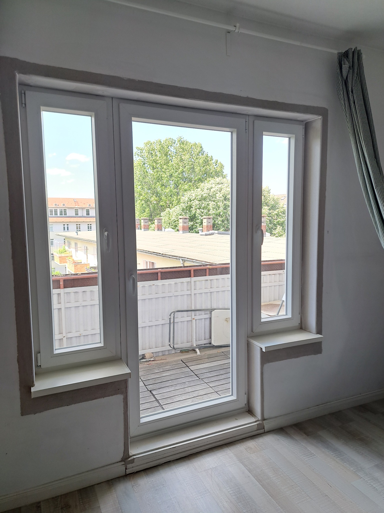 Białe okno balkonowe z widokiem na balkon i zieleń. Świeżo zamontowane, z widocznymi śladami po montażu na ścianie. Podłoga z paneli. Fragment zasłony w tle.