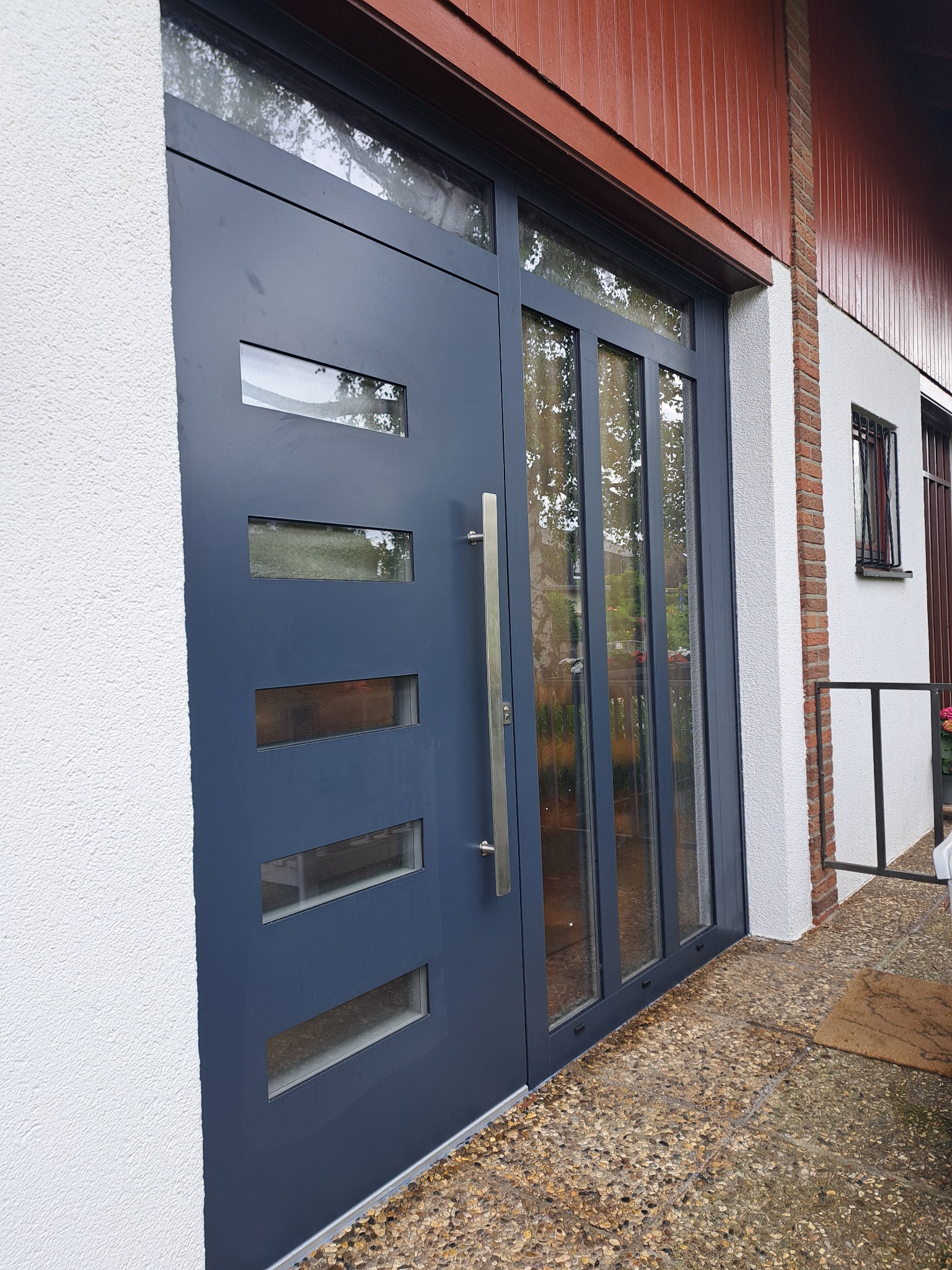 Nowoczesne, szare drzwi zewnętrzne z przeszkleniami i długim, pionowym uchwytem, zamontowane w białej elewacji budynku z brązowym drewnianym wykończeniem dachu.