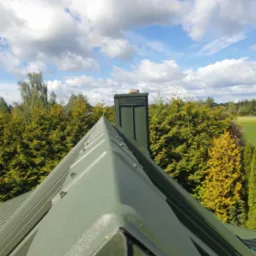 Szczegółowe ujęcie nowego, zielonego dachu blaszanego z kominem na tle błękitnego nieba z chmurami i zieleni drzew. Widok z góry ukazuje symetrię i detale konstrukcji.