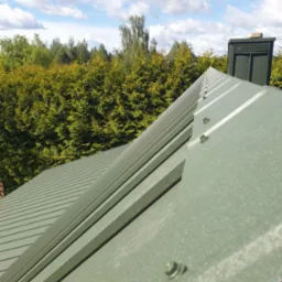 Zielony dach z blachy trapezowej, widok z góry na tle drzew i błękitnego nieba z chmurami. Widoczne detale łączeń i komin wentylacyjny.