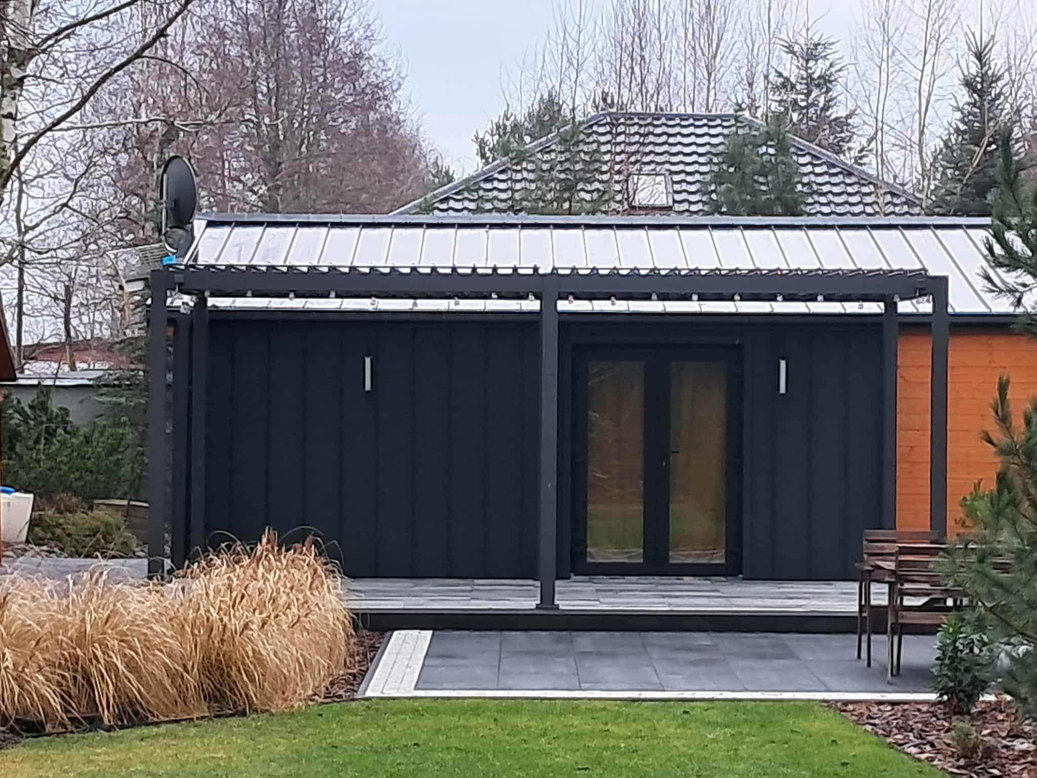 Nowoczesny budynek z ciemną elewacją i metalowym zadaszeniem. Widoczny fragment dachu z dachówką, zadbany trawnik i ozdobne trawy. Antena satelitarna na dachu.