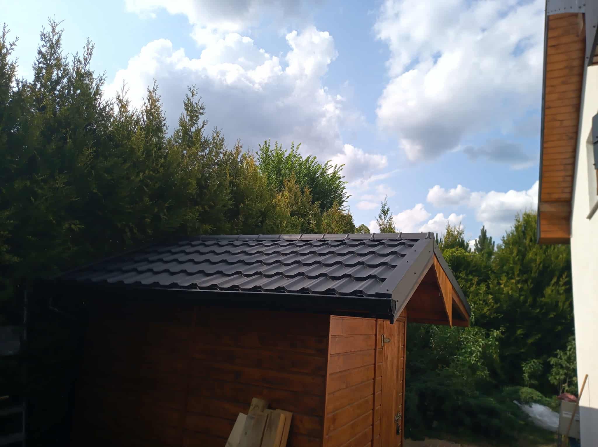 Drewniany domek narzędziowy z czarnym, metalowym dachem modułowym na tle błękitnego nieba i zieleni drzew. Widoczne deski przed domkiem.