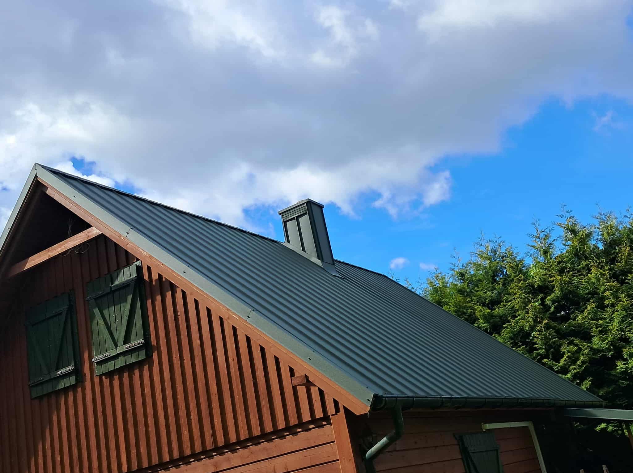 Dom z nowym, ciemnym dachem z blachy trapezowej, brązowe deski elewacyjne i zielone okiennice, na tle błękitnego nieba z chmurami i zielonych drzew.