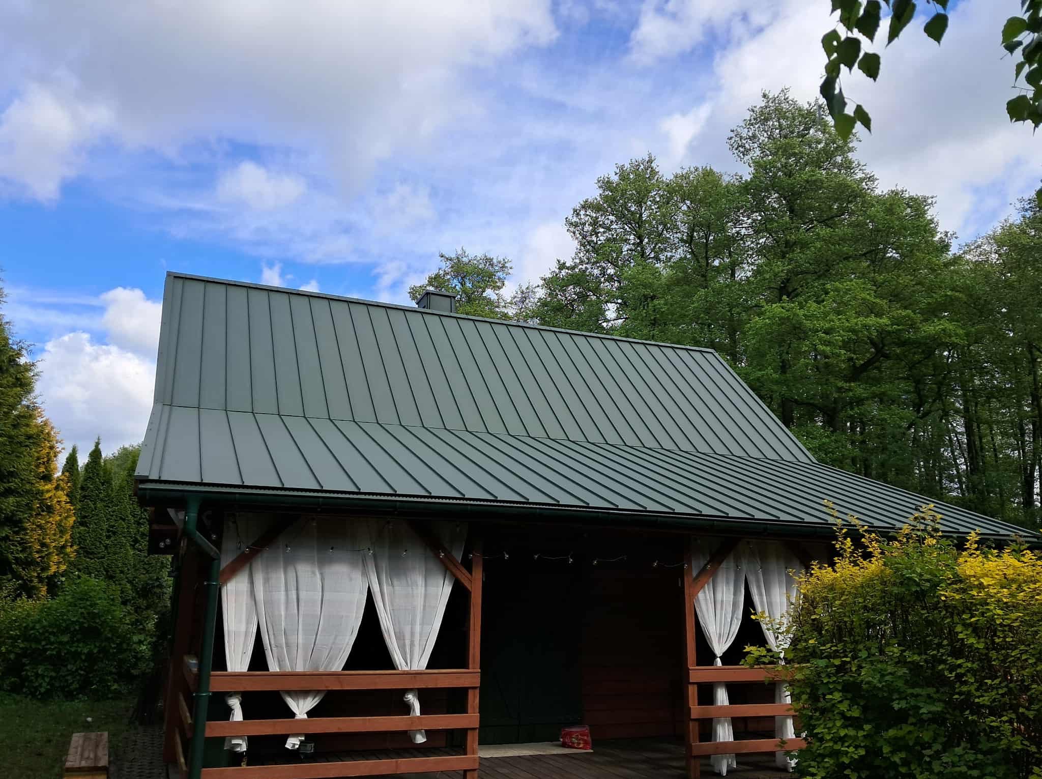 Zadaszona altana z zielonym dachem z blachy trapezowej i białymi zasłonami, otoczona zielenią. Nowoczesne krycie dachu na tle błękitnego nieba.