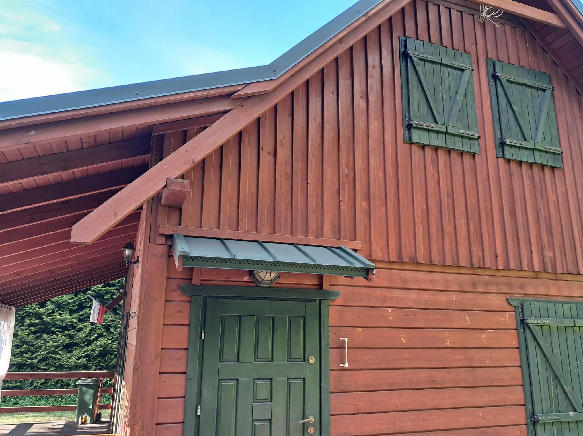 Drewniany dom z ciemnozielonymi okiennicami i dachem. Widoczna część dachu i elewacji. Na balkonie flaga Polski.