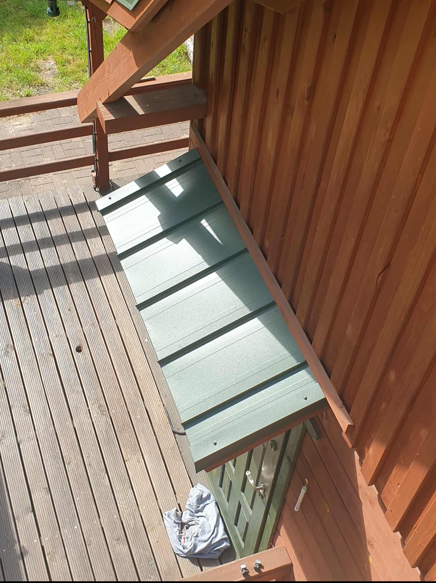 Z bliska: fragment dachu nad wejściem do domku, wykonany z blachy trapezowej w odcieniach zieleni. Widoczna drewniana konstrukcja i fragment drewnianego tarasu.