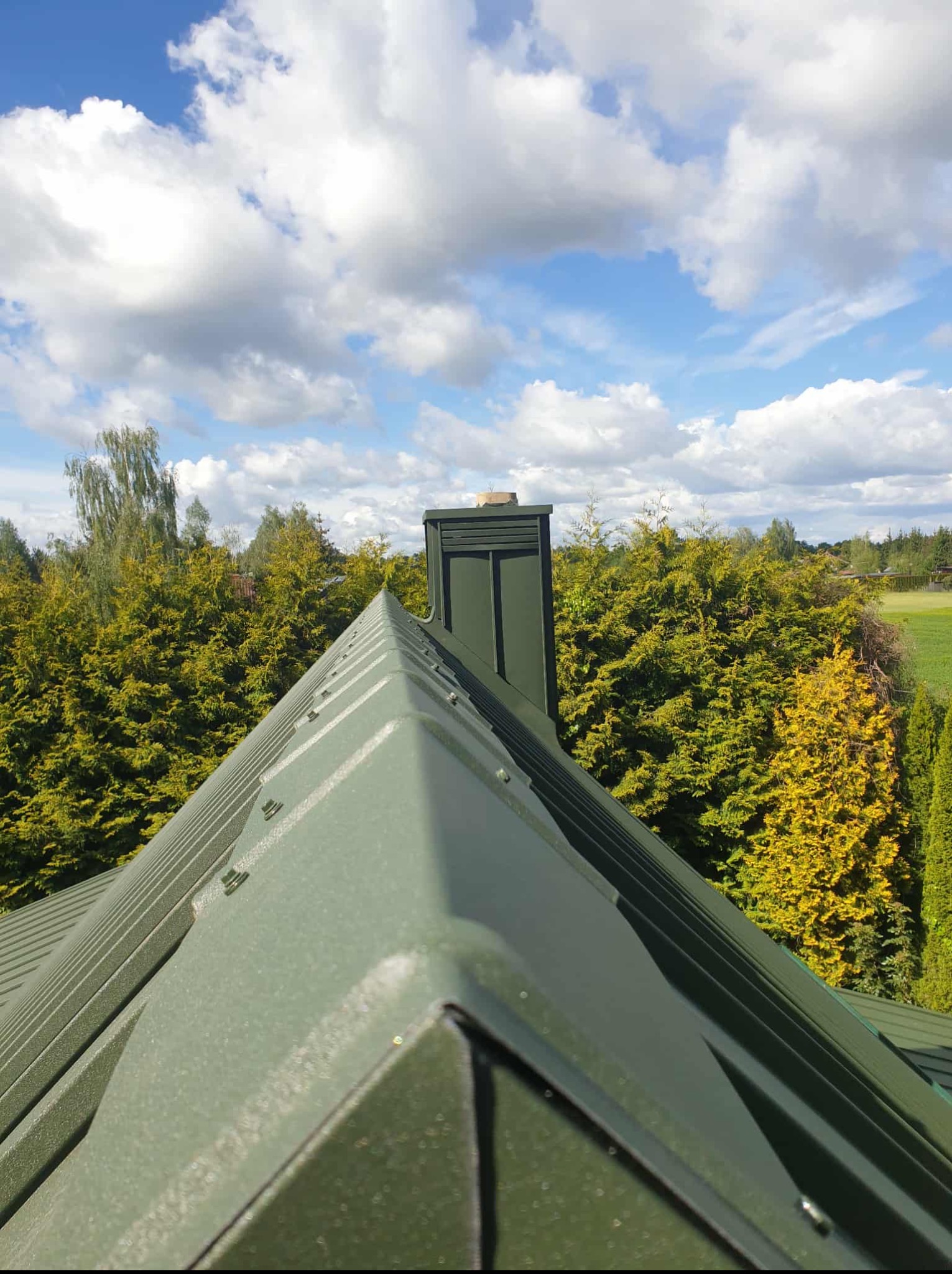 Szczegółowe ujęcie nowego, zielonego dachu blaszanego z kominem na tle błękitnego nieba z chmurami i zieleni drzew. Widok z góry ukazuje symetrię i detale konstrukcji.