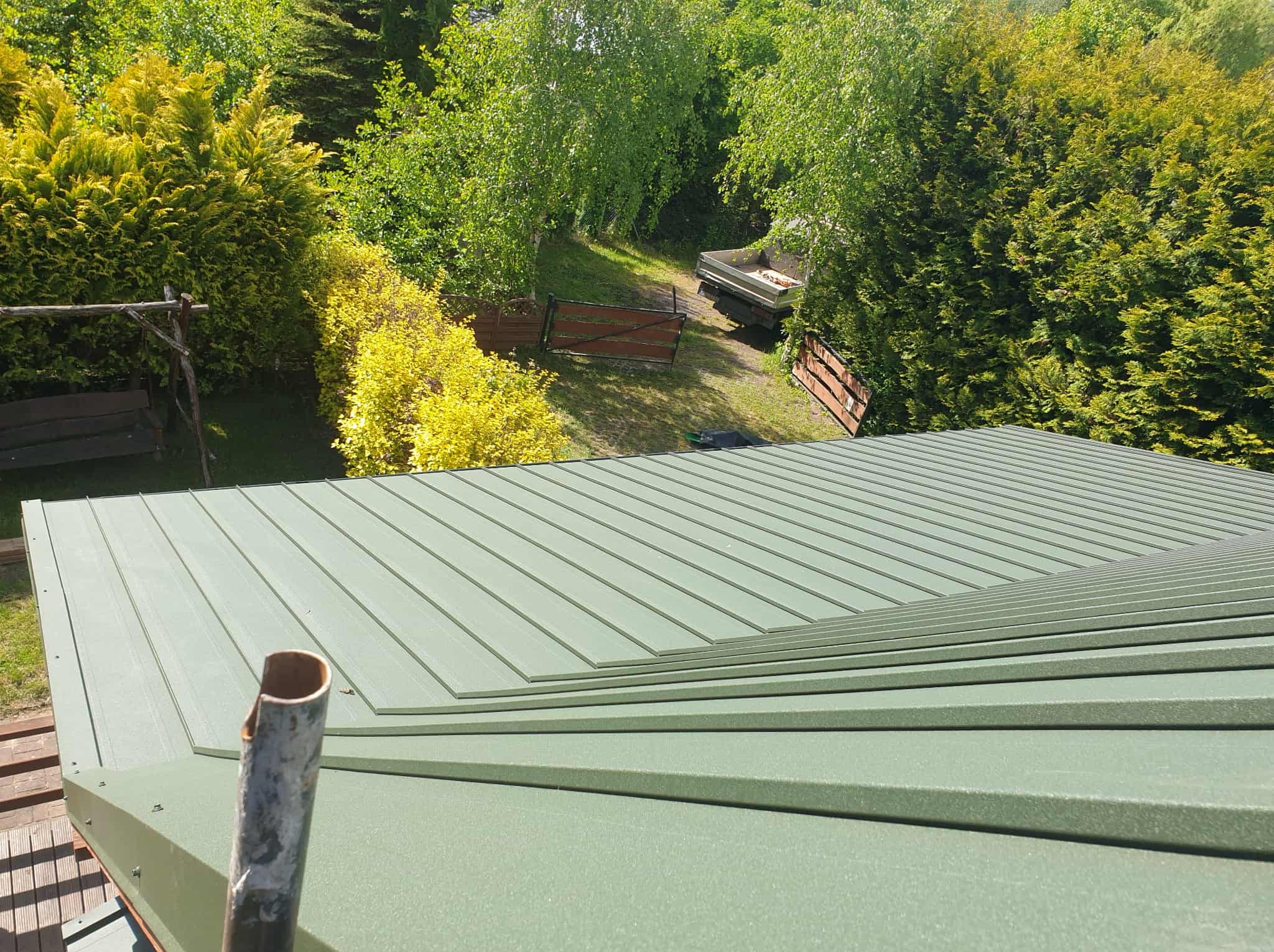 Zielony dach z blachy trapezowej w słoneczny dzień, z widokiem na bujną zieleń drzew i krzewów w tle. Fragment rury spustowej w lewym dolnym rogu kadru.