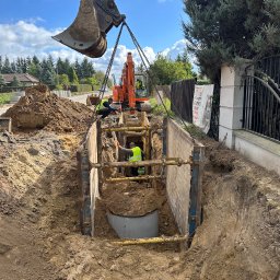 ProHouse - Wykop zabezpieczony szalunkiem stalowym. Koparka z łyżką opuszcza betonowy krąg do wykopu. Dwóch pracowników w żółtych kamizelkach nadzoruje prace.