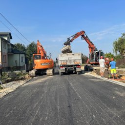 ProHouse - Prace ziemne w Jaroszewicach: Dwie koparki Doosan i Terex ładują urobek na ciężarówkę LU42SRG. Widoczni pracownicy i niwelator. Asfaltowa droga po prawej.