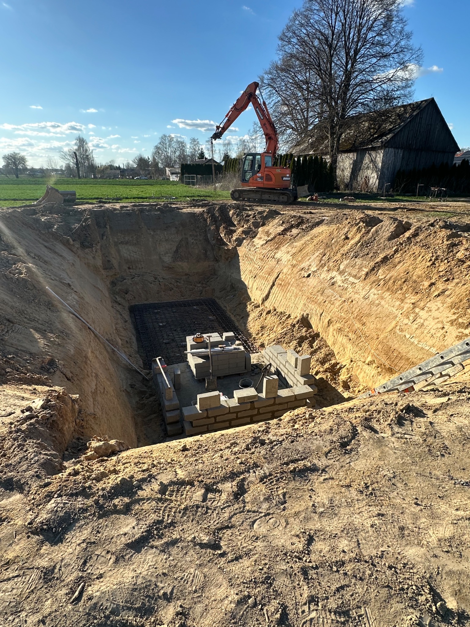 Wykop pod szambo z widocznym zbrojeniem i częściowo wymurowaną konstrukcją. Na górze koparka. Słoneczny dzień, wiejski krajobraz w tle.