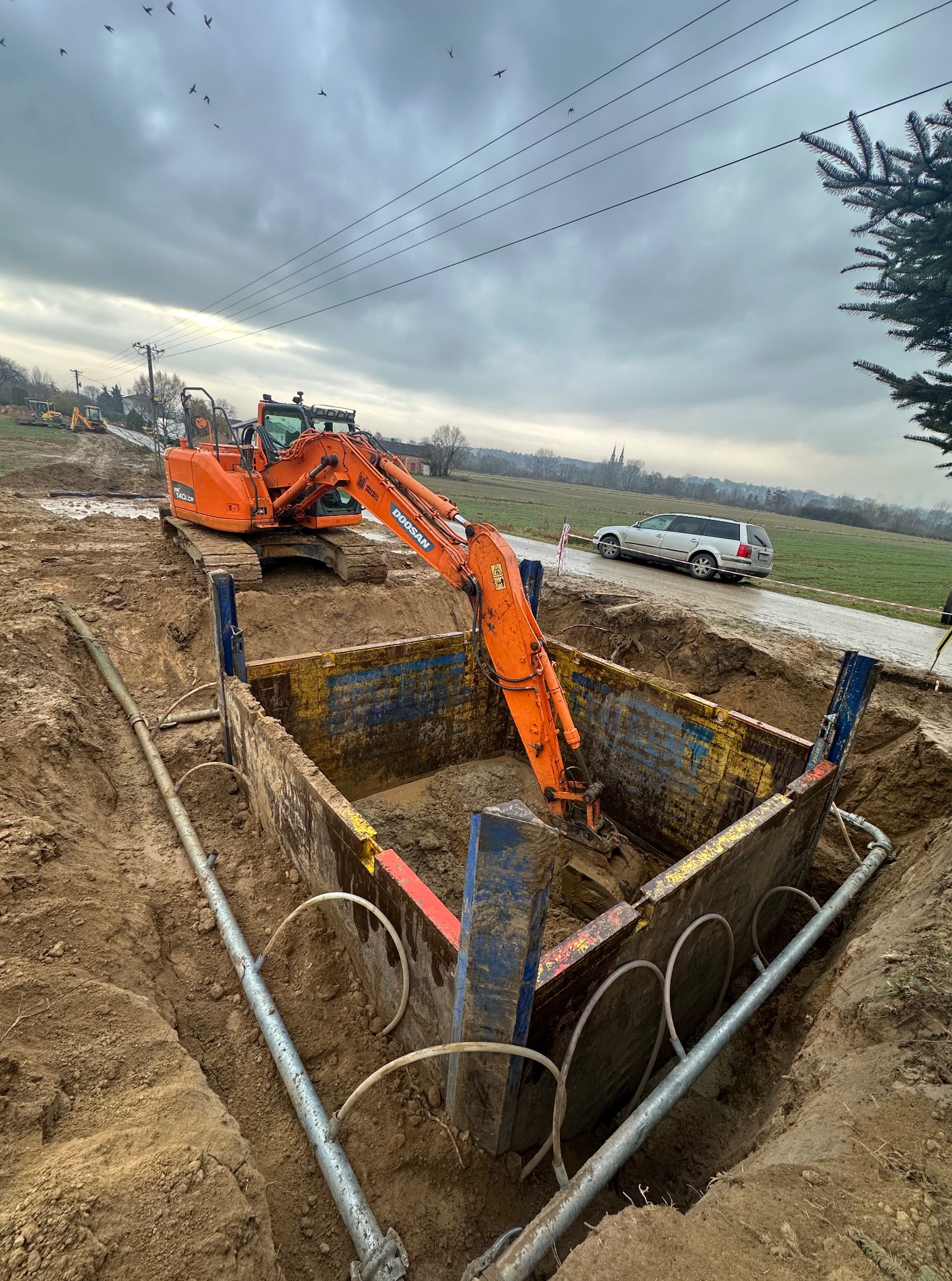 Pomarańczowa koparka Doosan w wykopie zabezpieczonym szalunkiem. W tle widoczne pole, drzewa, samochód i pochmurne niebo z kluczem ptaków.