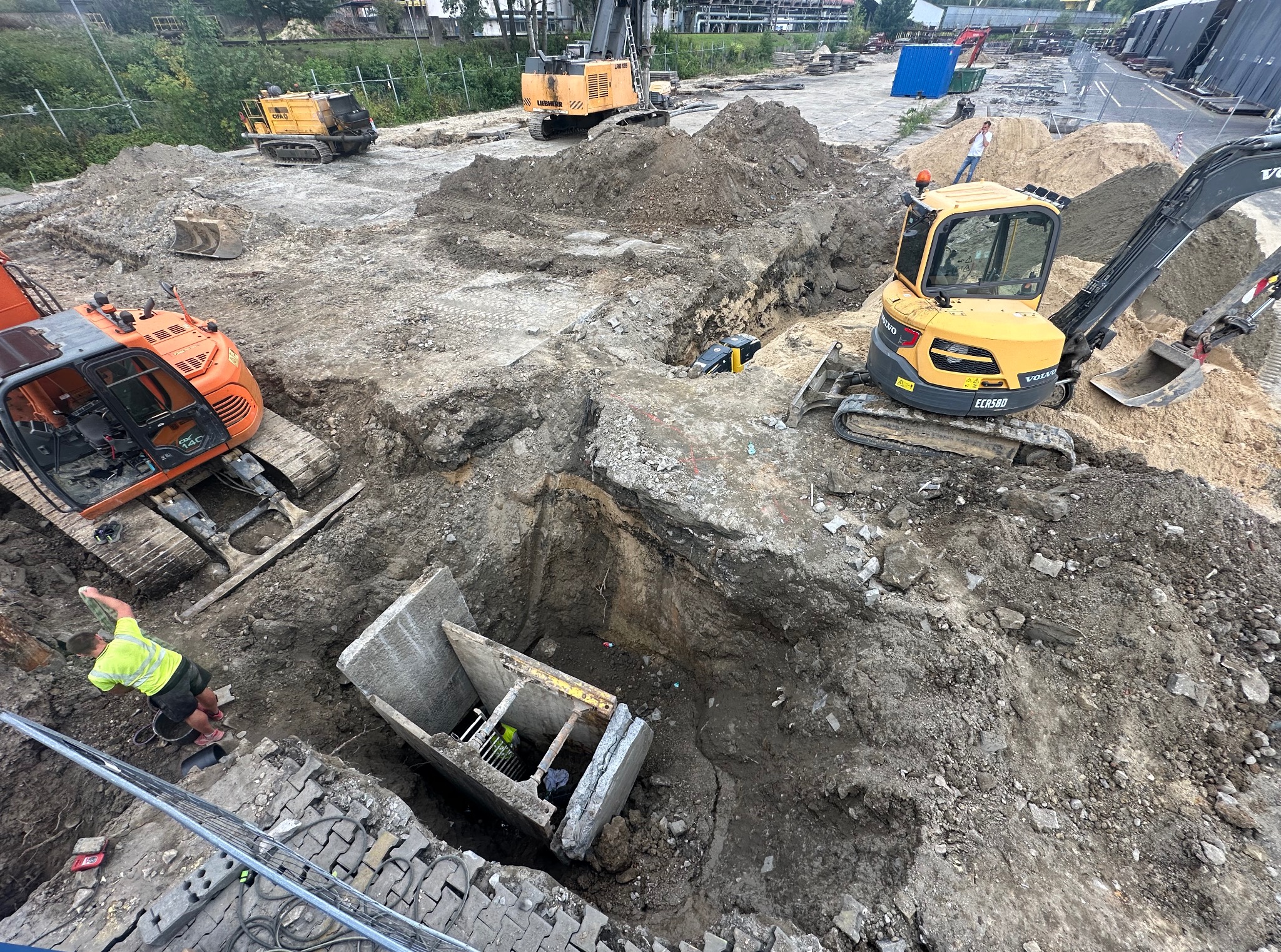 Głęboki wykop z betonowymi elementami, praca wre. Żółta koparka Volvo ECR58D i pomarańczowa maszyna Hitachi w akcji. Pracownicy w dole i na górze. Wykop pod szambo.