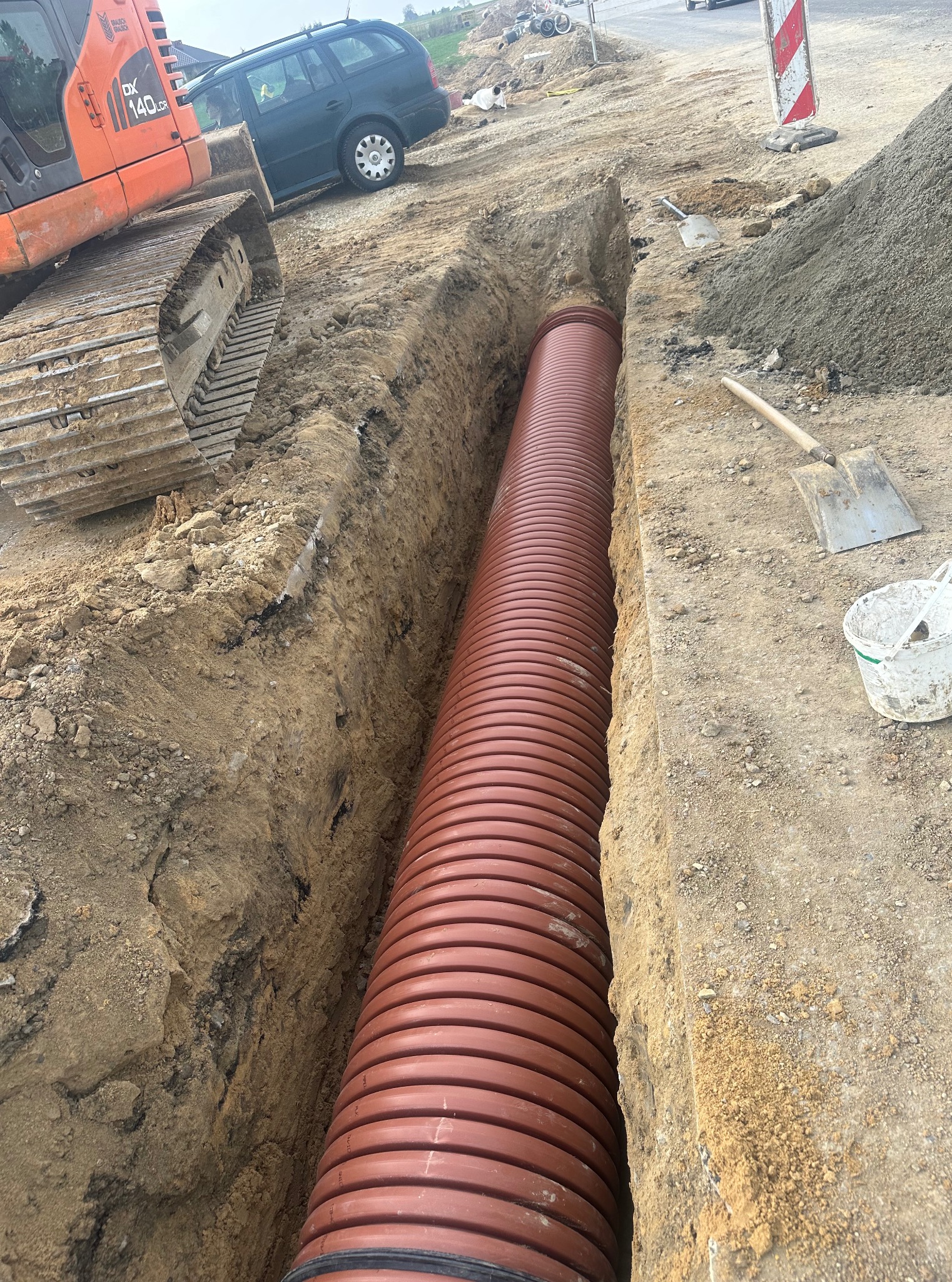 Długi, brązowy rurociąg z tworzywa w wykopie ziemnym, obok koparka i sterta piasku. Widoczne narzędzia: łopata i wiadro. Prace ziemne w toku.