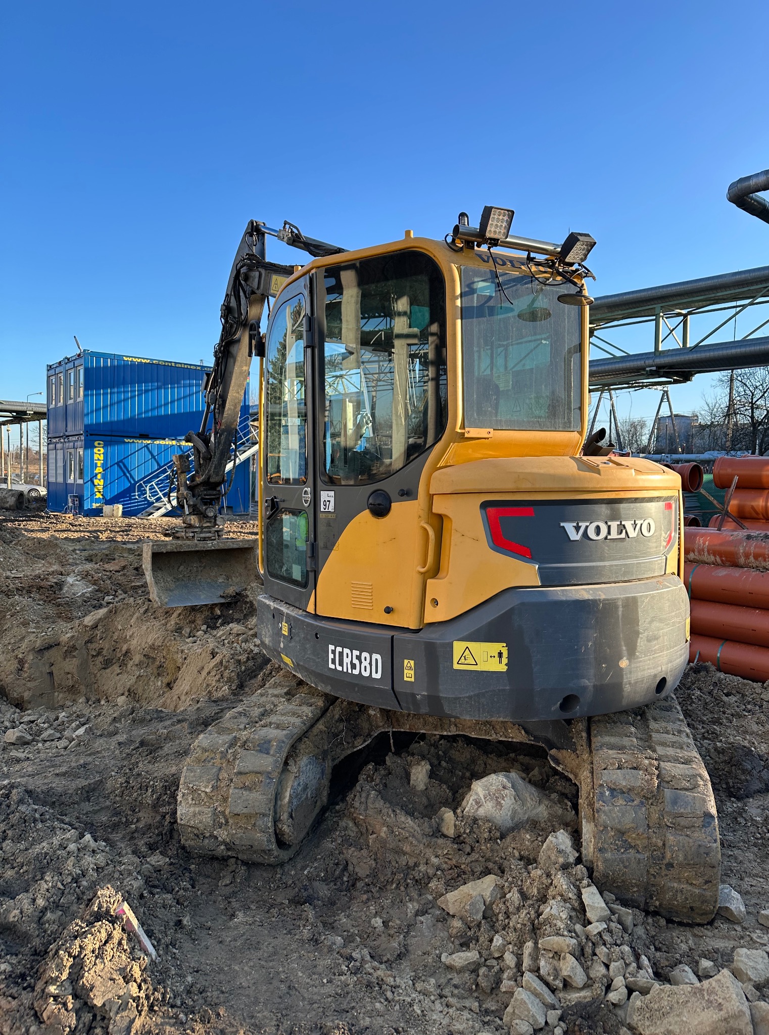 Żółta koparka Volvo ECR58D na placu budowy, z wykopem w ziemi, rurami i kontenerami w tle, pod błękitnym niebem. Widok z przodu na maszynę gotową do pracy.