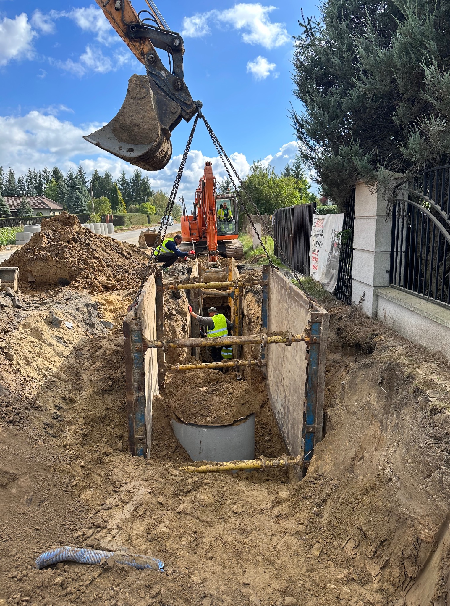 Wykop zabezpieczony szalunkiem stalowym. Koparka z łyżką opuszcza betonowy krąg do wykopu. Dwóch pracowników w żółtych kamizelkach nadzoruje prace.