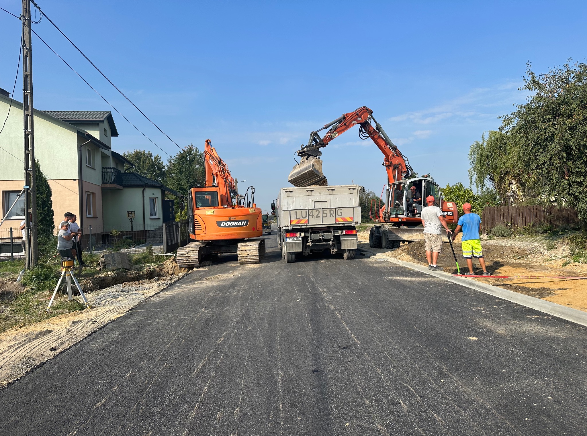Prace ziemne w Jaroszewicach: Dwie koparki Doosan i Terex ładują urobek na ciężarówkę LU42SRG. Widoczni pracownicy i niwelator. Asfaltowa droga po prawej.