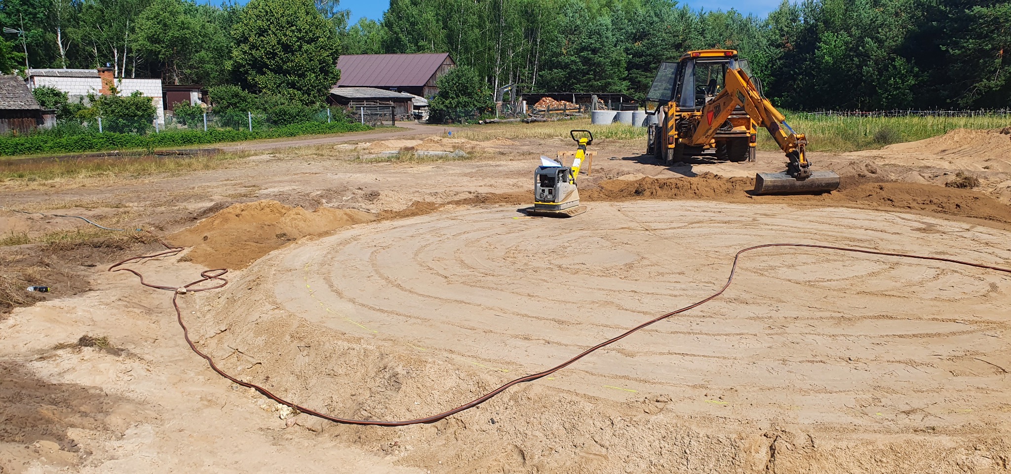 Żółta koparko-ładowarka i zagęszczarka w trakcie wyrównywania terenu pod okrągły zbiornik. Widoczne ślady opon i przewód zasilający. W tle drzewa i zabudowania.