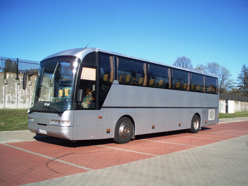 Srebrny autokar turystyczny na parkingu z kostki, widok z boku, błękitne niebo w tle.