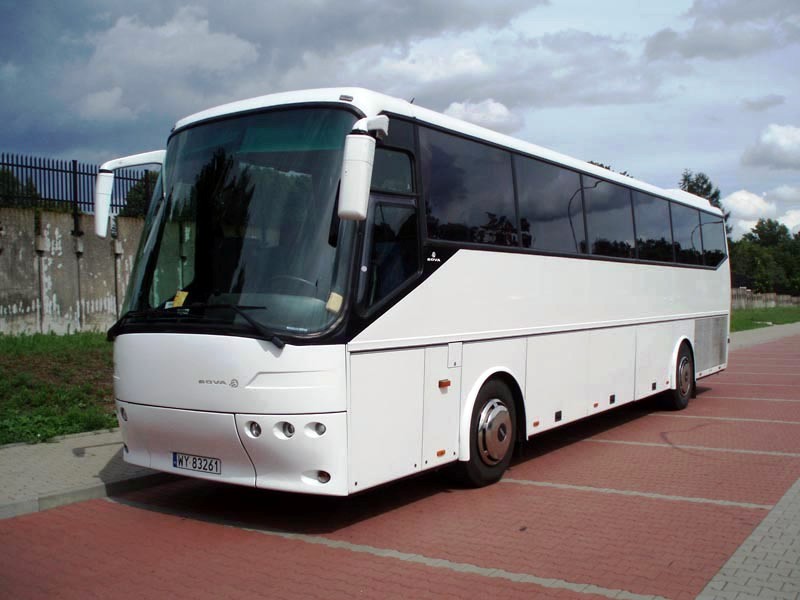 Biały autokar marki Bova na czerwonym brukowanym parkingu z metalowym ogrodzeniem w tle.