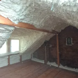 Poddasze po ociepleniu pianką PUR. Widoczne drewniane belki, okno i fragment ściany z cegły. Pianka o fakturze chropowatej, pokrywa skośny sufit i fragmenty ścian.