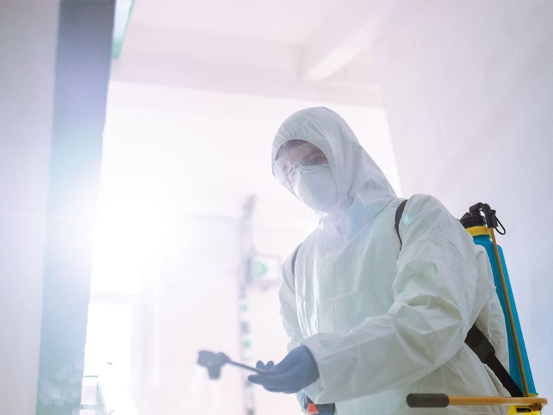 Pracownik w kombinezonie ochronnym, z maską i opryskiwaczem, wykonuje zabieg w jasnym pomieszczeniu. Widoczny opryskiwacz plecakowy i lanca opryskowa.