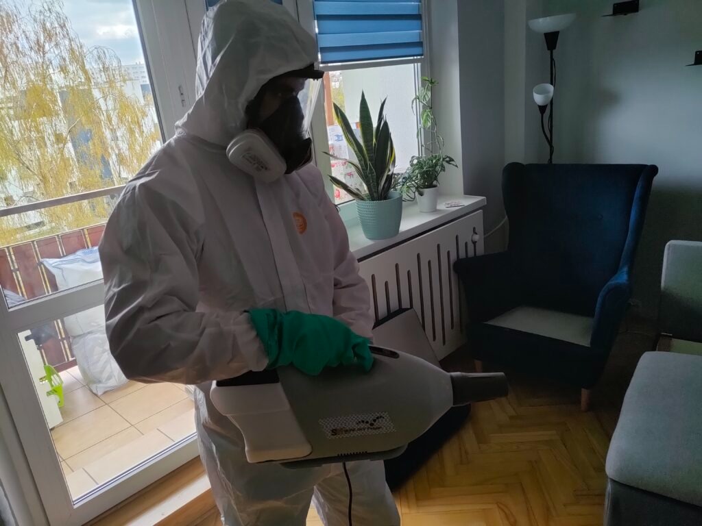 Osoba w kombinezonie ochronnym, masce i zielonych rękawicach dezynfekuje pomieszczenie za pomocą zamgławiacza. Widoczne meble i roślina doniczkowa.