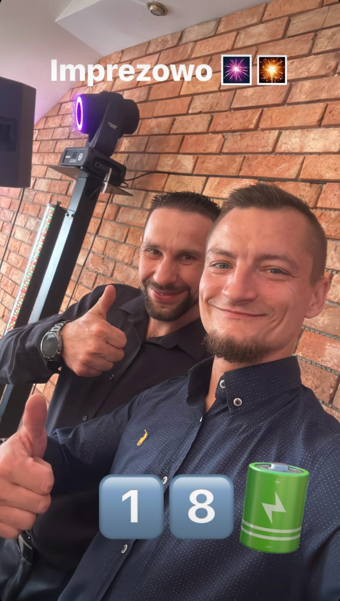Uśmiechnięci mężczyźni na tle ceglanej ściany z oświetleniem imprezowym. Jeden z nich pokazuje kciuk w górę. Widoczne elementy dekoracyjne i napis 'Imprezowo'.