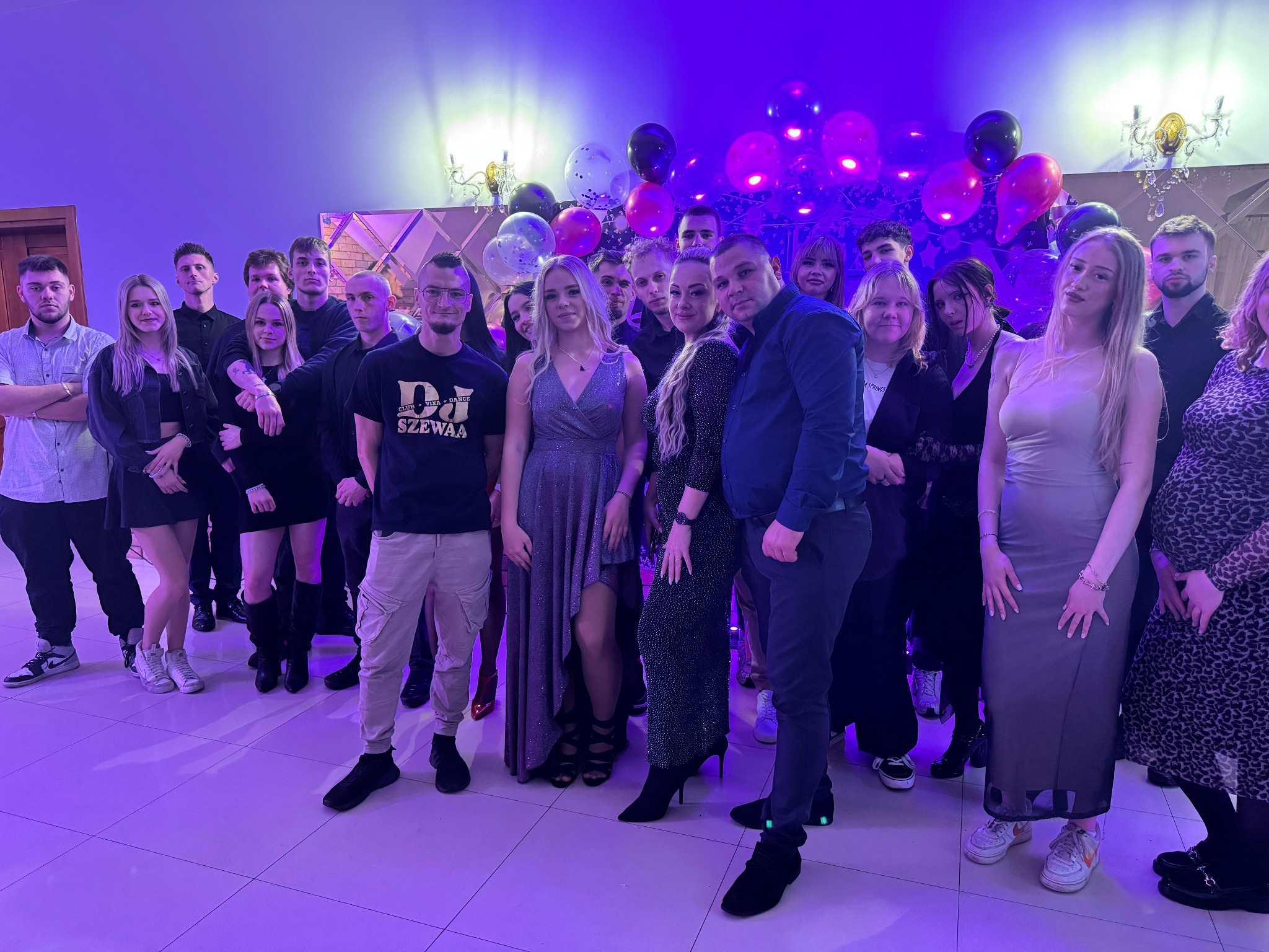 Grupowe zdjęcie zespołu muzycznego 'DJ SZEWAA' i gości na tle dekoracji z balonów podczas imprezy oświetlonej fioletowym światłem.