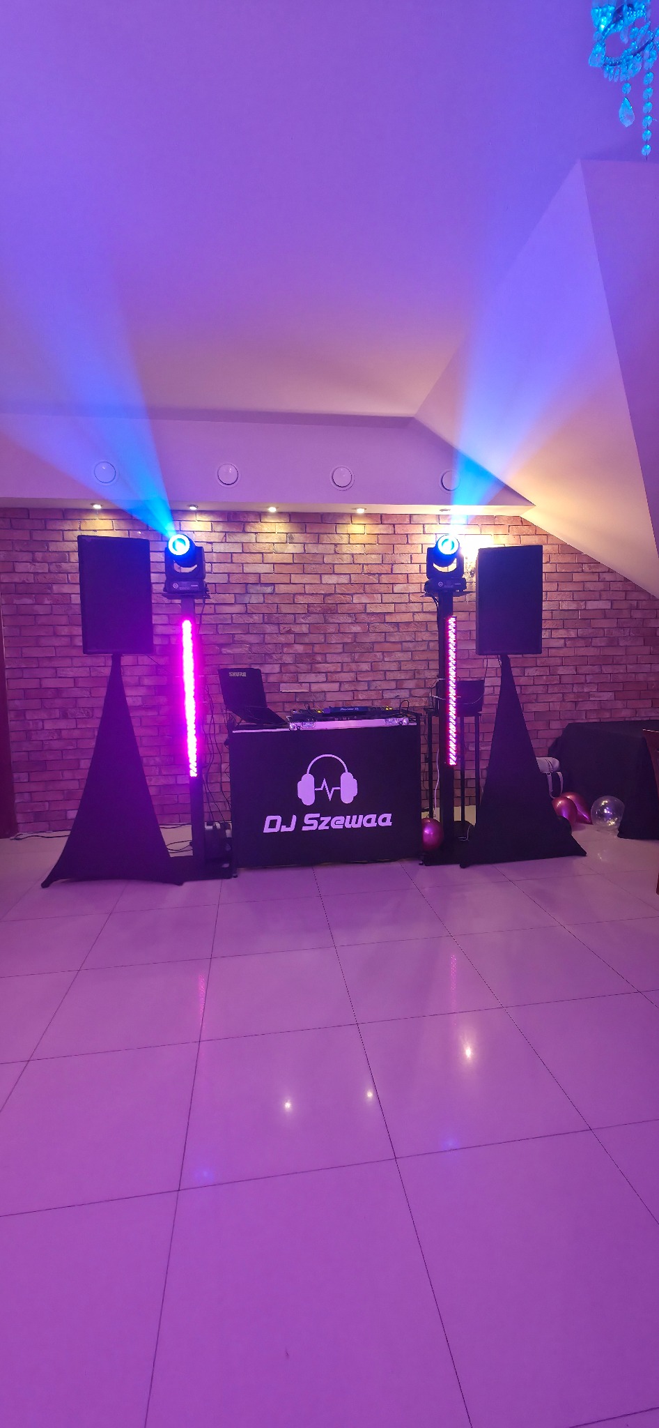 Stanowisko DJ'a 'DJ Szewaa' z nagłośnieniem i oświetleniem na tle ceglanej ściany, oświetlone fioletowym światłem. Widoczne kolumny, reflektory i konsoleta.