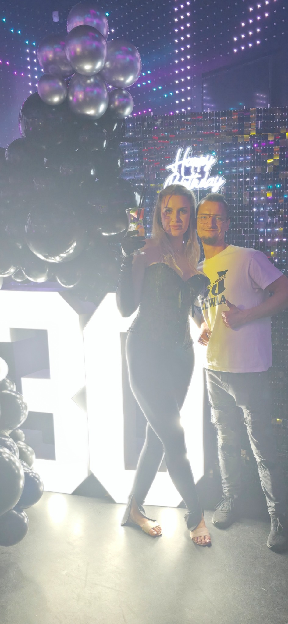 Uśmiechnięta para pozuje na tle dekoracji urodzinowych: czarne balony, błyszcząca ściana i podświetlony napis '30'. Kobieta trzyma drinka.