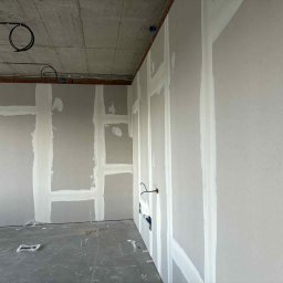 EL-KRAFT Usługi Ogólnobudowlane Tomasz Krefta - Wnętrze w trakcie prac: ściany z płyt, widoczne łączenia wzmocnione taśmą i pokryte masą szpachlową. Na suficie widoczne instalacje elektryczne. Surowy stan wykończenia.