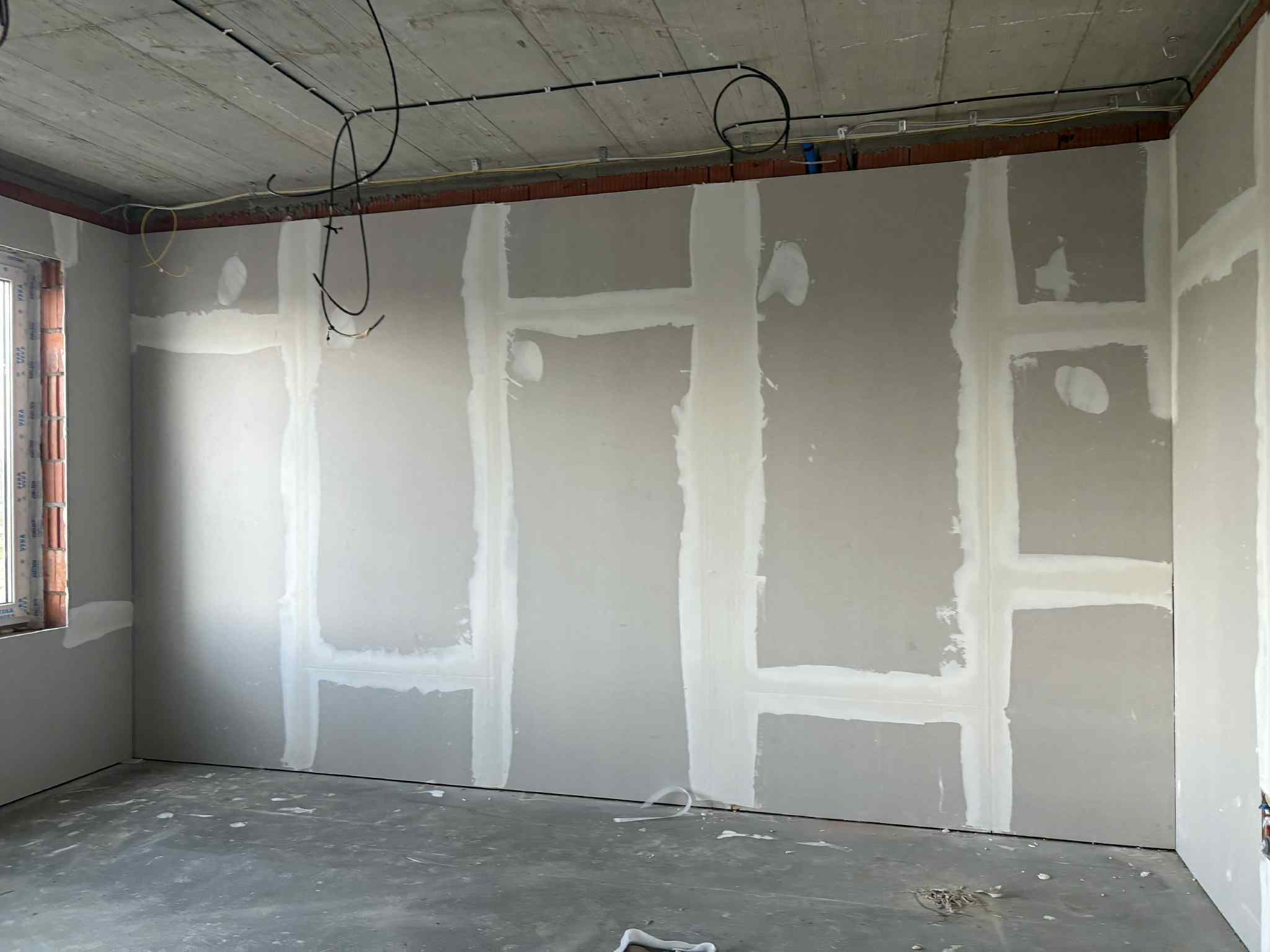 Wnętrze w trakcie prac: ściany z płyt gipsowo-kartonowych, widoczne łączenia szpachlowane. Na suficie instalacja elektryczna. Surowy betonowy sufit i podłoga.