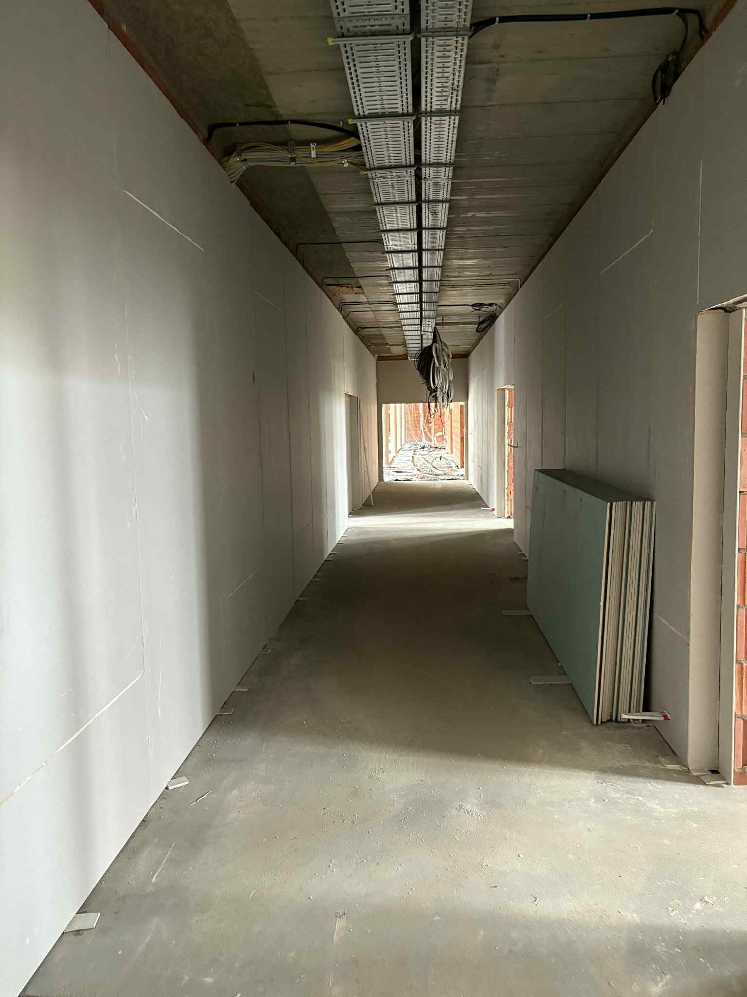 Korytarz w trakcie wykańczania: zamontowane płyty karton-gips na ścianach, widoczne kable pod sufitem i stos płyt przy ścianie. Surowy beton na podłodze.