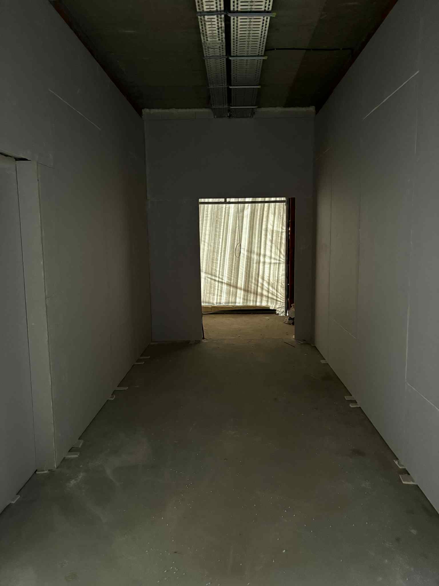 Korytarz w trakcie prac wykończeniowych, ściany pokryte szarą płytą. Widoczne profile i instalacje pod sufitem. W głębi osłonięte wejście.