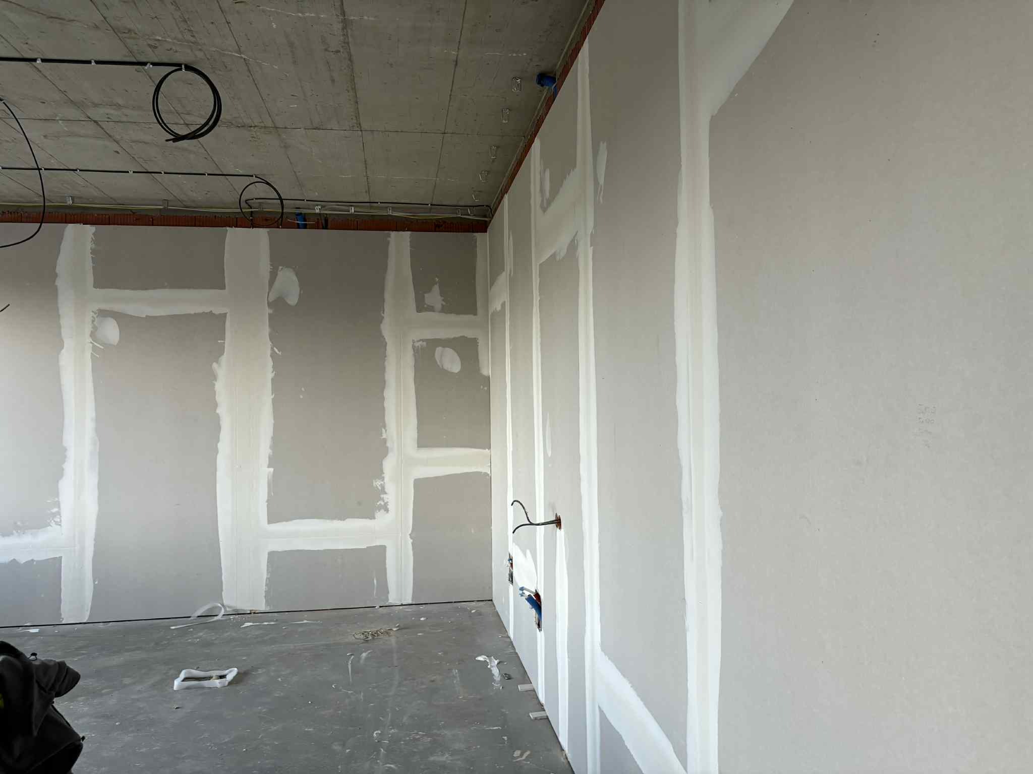 Wnętrze w trakcie prac: ściany z płyt, widoczne łączenia wzmocnione taśmą i pokryte masą szpachlową. Na suficie widoczne instalacje elektryczne. Surowy stan wykończenia.