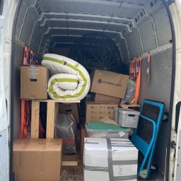 Mrówka Trans - Wnętrze białego busa transportowego wypełnione kartonami (IKEA, Samsung), zwiniętym materacem, niebieskim wózkiem i zabezpieczone pomarańczowymi pasami, gotowe do przeprowadzki.
