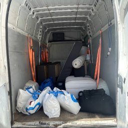 Mrówka Trans - Wnętrze białego samochodu dostawczego, wypełnione po brzegi przedmiotami: białymi torbami z niebieskimi uchwytami, zwiniętym dywanem, poduszką i pudłami, zabezpieczonymi pomarańczowymi pasami.
