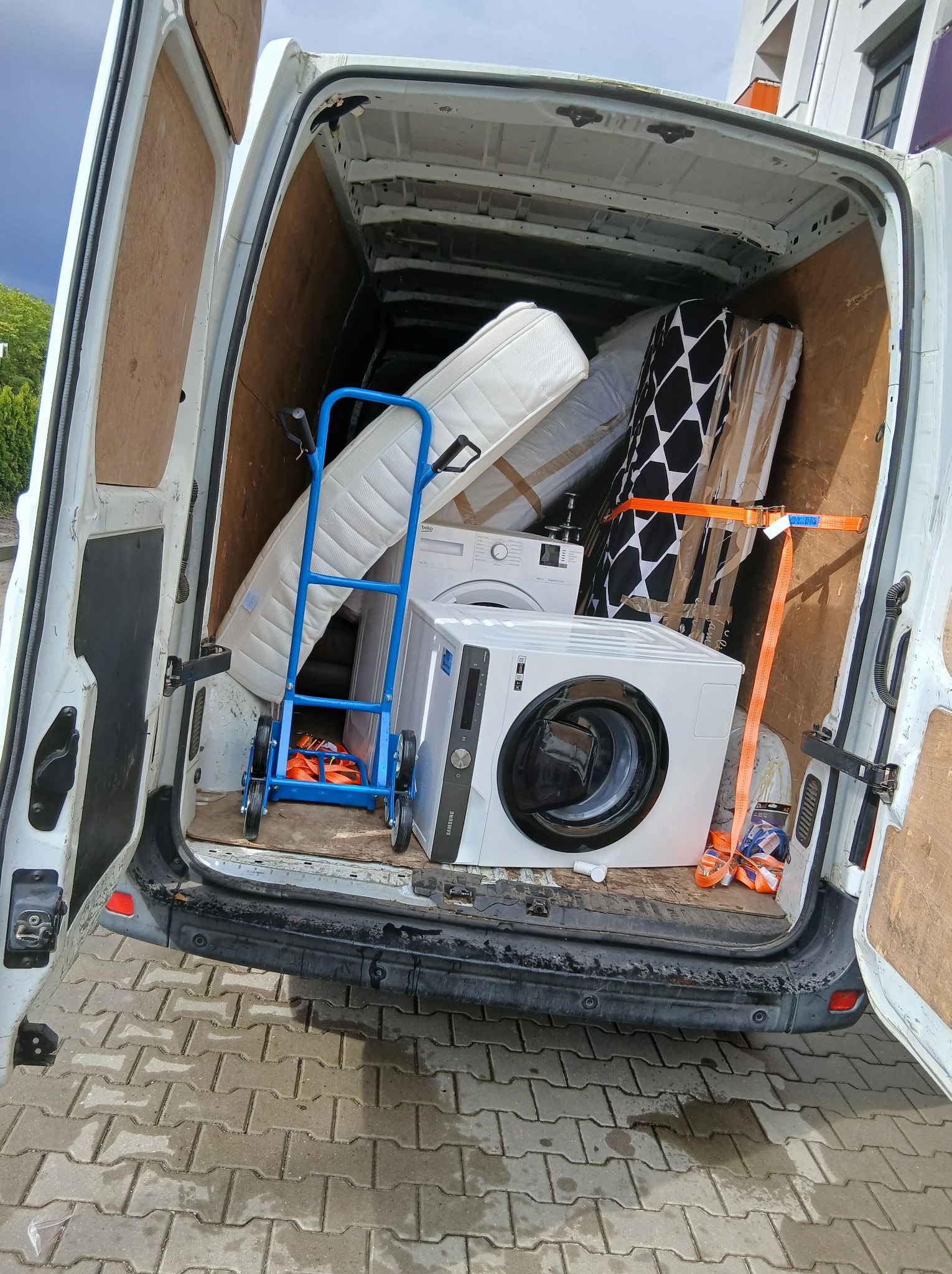 Wnętrze załadowanego busa z pralkami, materacami i wózkiem transportowym, zabezpieczone pasami. Widok z tyłu pojazdu na kostkę brukową.