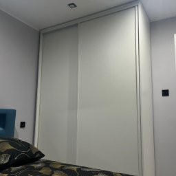 We made it Tomasz Białek - Biała szafa przesuwna z lustrzanymi drzwiami, wkomponowana w jasne wnętrze sypialni, widok z perspektywy łóżka z wzorzystą pościelą.