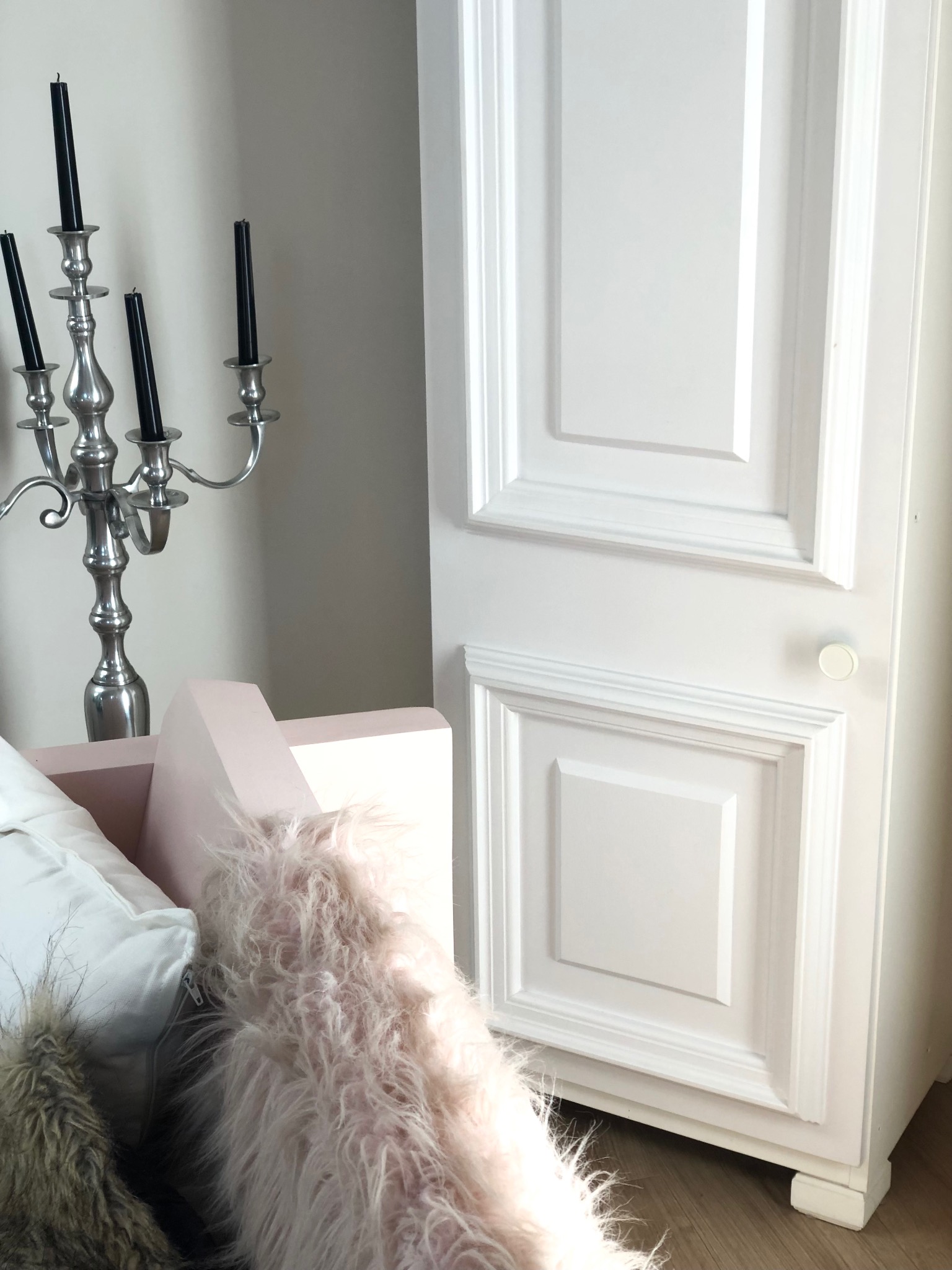 Biała, panelowa szafka z okrągłym uchwytem, obok srebrny świecznik z czarnymi świecami i fragment różowego fotela z futrzanym pledem w tle.