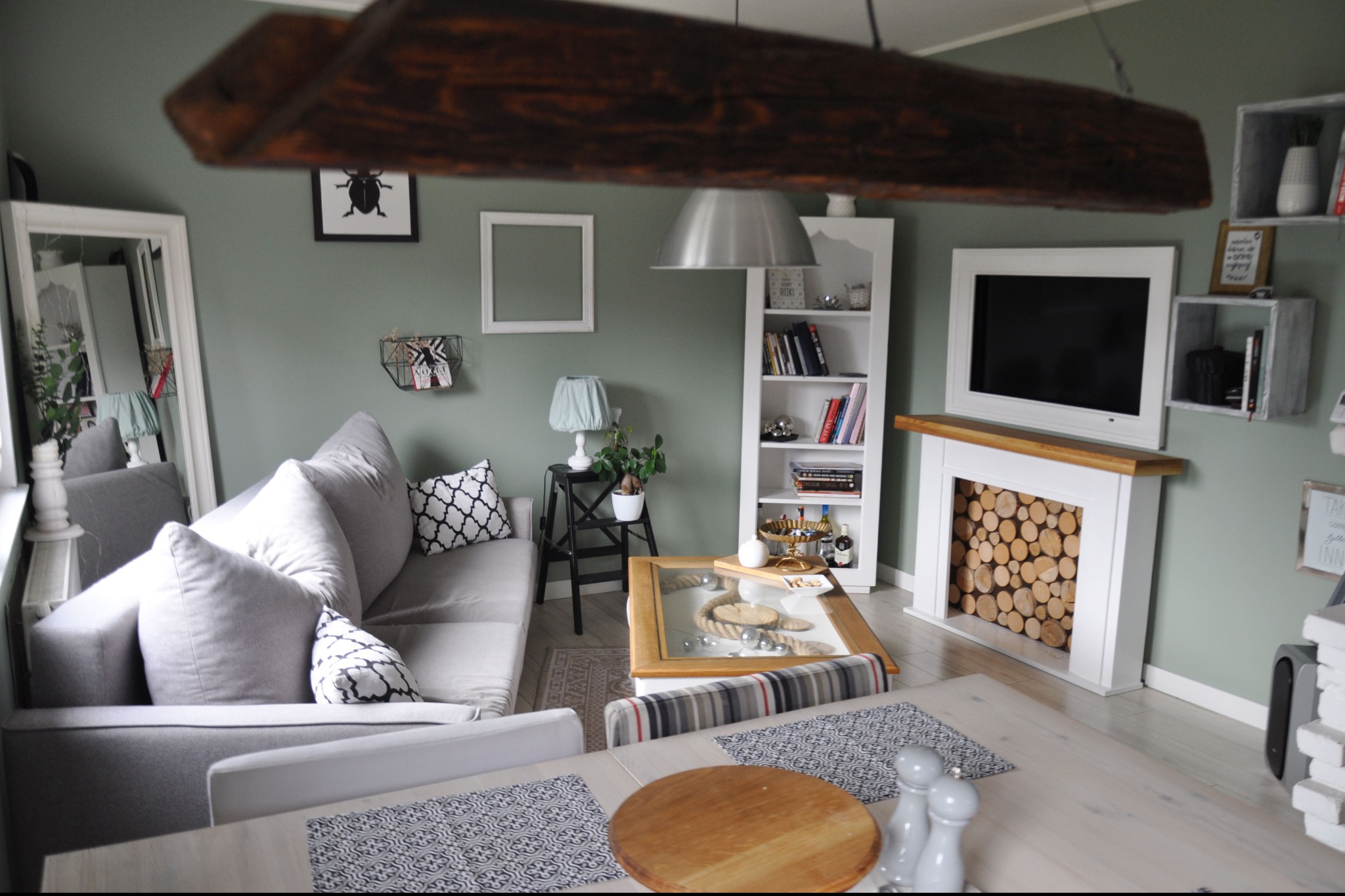 Jasnoszary salon z drewnianą belką sufitową, kominkiem z drewnem i szarą sofą. Widoczne półki na ścianie i lustro. Całość w stonowanej kolorystyce.