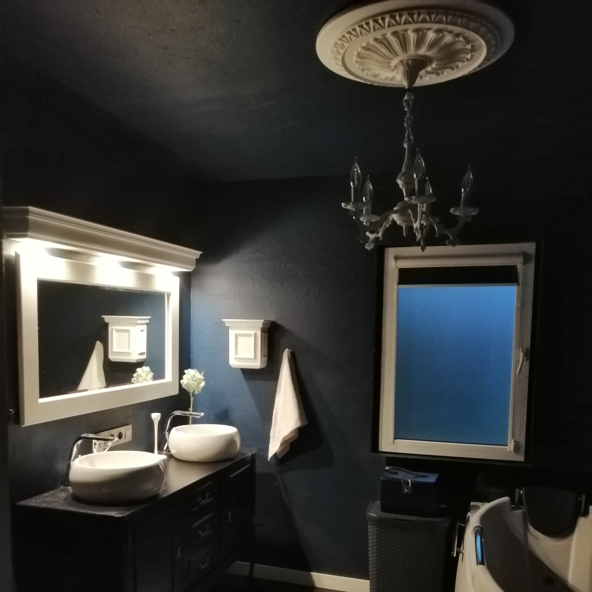 Elegancka łazienka w odcieniach granatu z białymi akcentami: umywalki, lustro z oświetleniem, okno z roletą i żyrandol. Nowoczesny wystrój wnętrza.