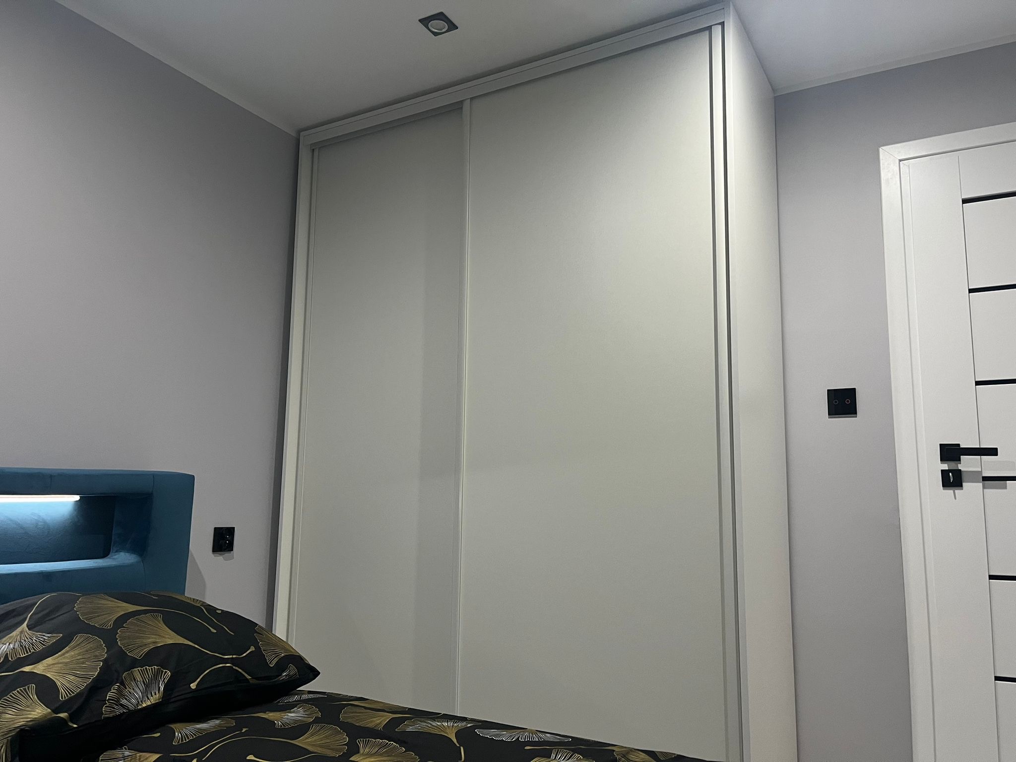 Biała szafa przesuwna z lustrzanymi drzwiami, wkomponowana w jasne wnętrze sypialni, widok z perspektywy łóżka z wzorzystą pościelą.