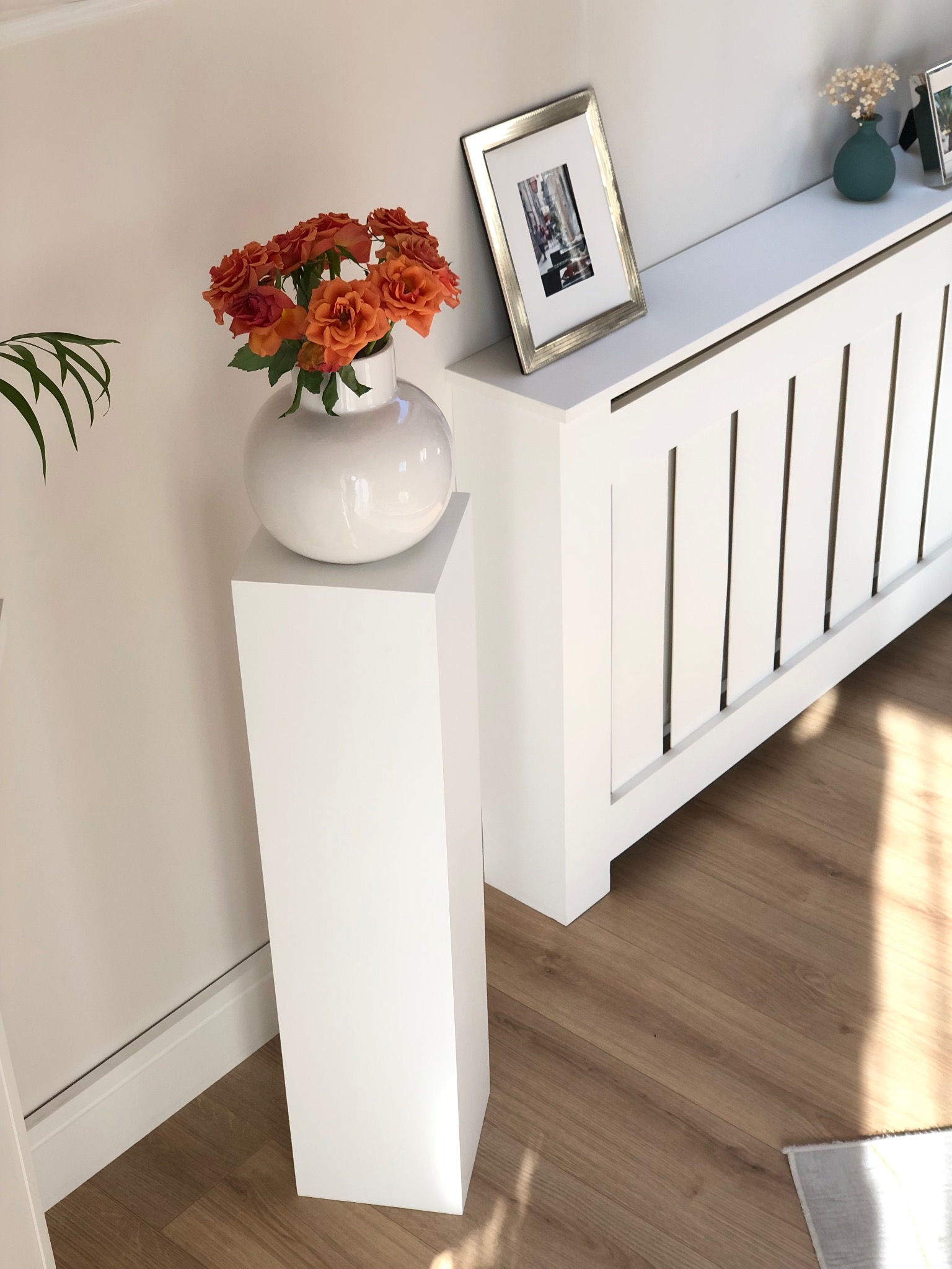 Biała zabudowa grzejnika z ramką i wazonem na blacie, obok kolumna z kwiatami. Minimalistyczny design wnętrza, jasne kolory, drewniana podłoga.