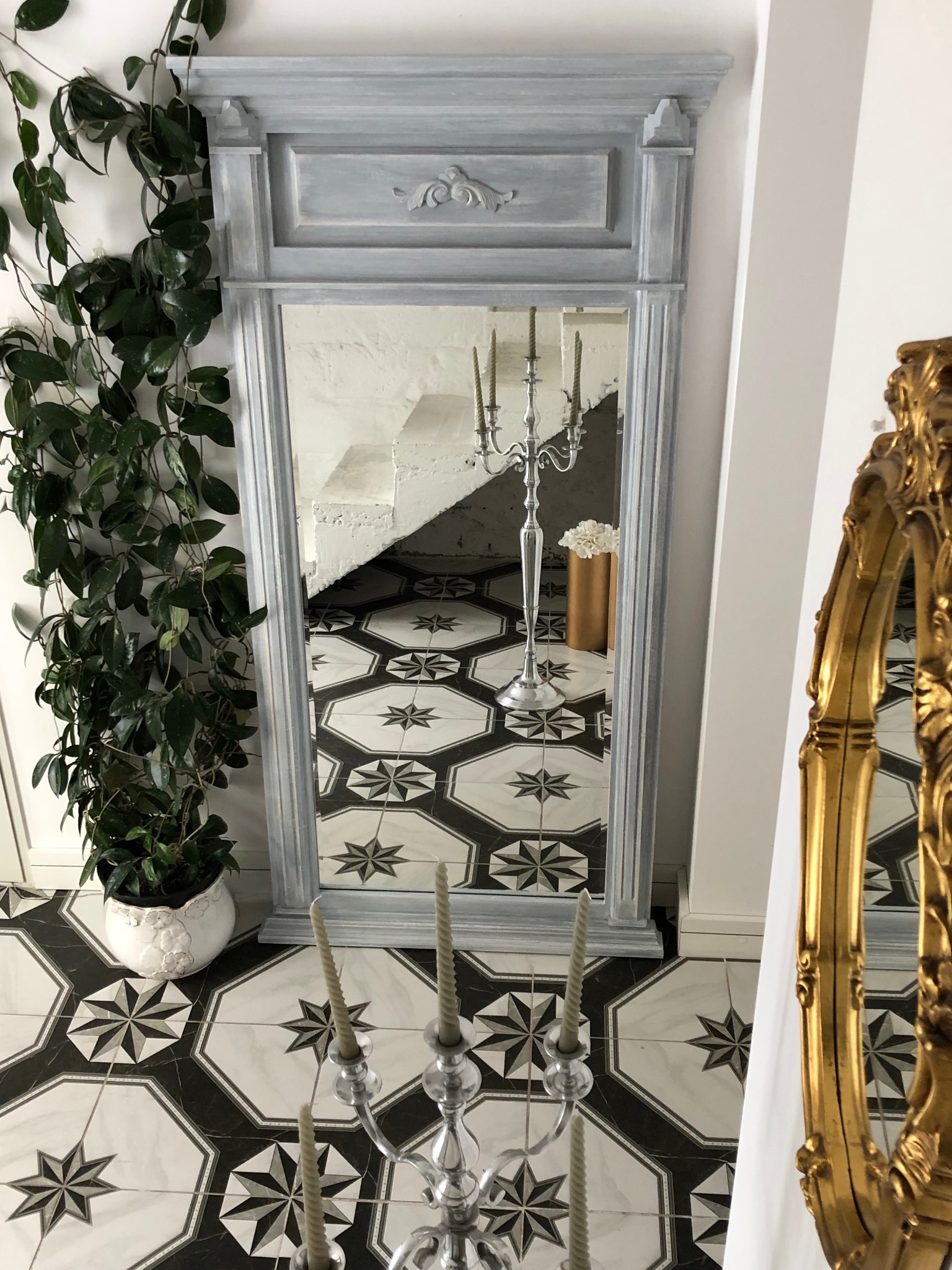 Stylowe lustro w szarej ramie z dekoracyjnymi elementami, odbijające schody i świecznik. Obok lustra roślina doniczkowa i fragment złotej ramy.