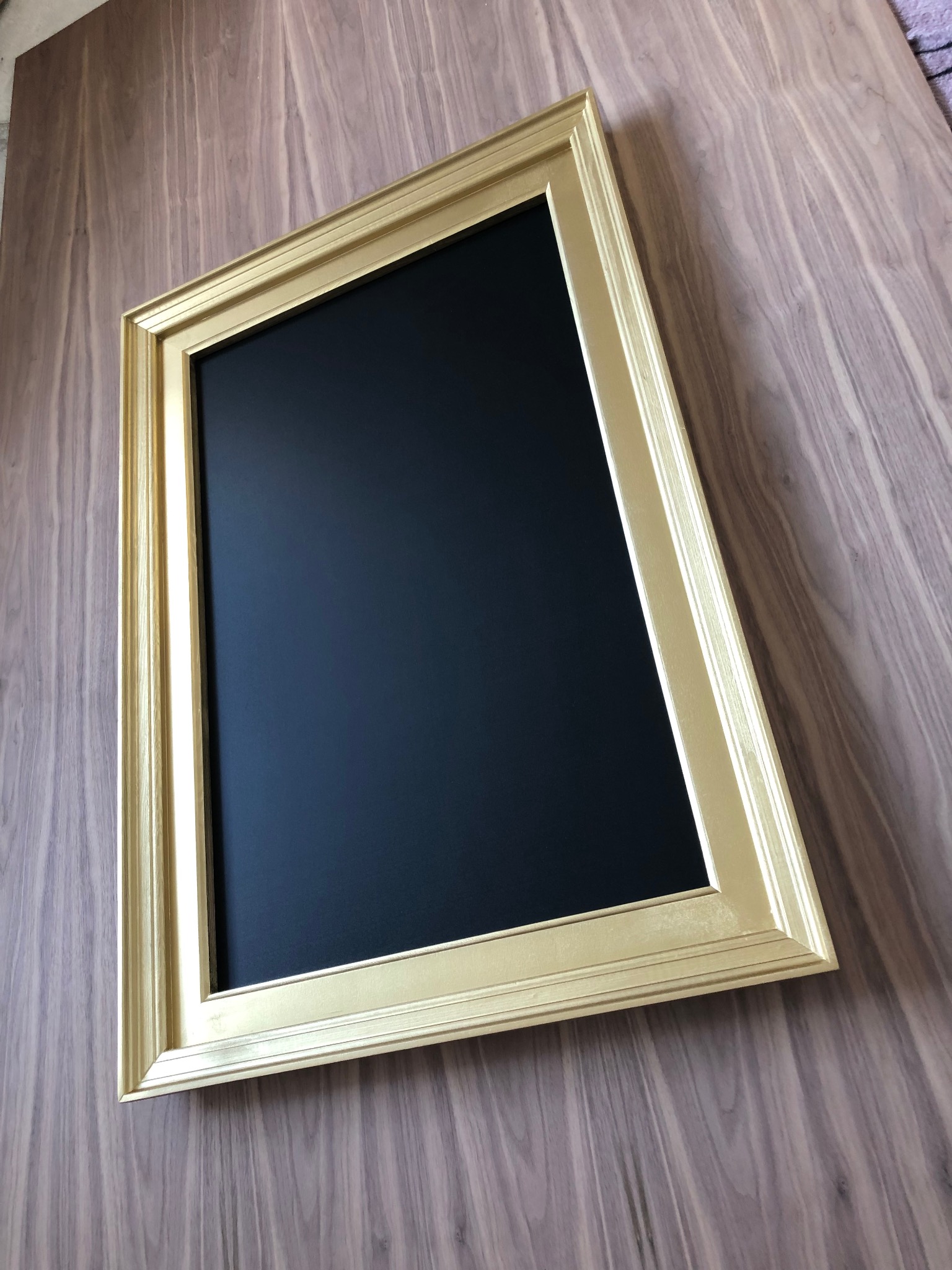 Złota rama lustra/obrazu na ścianie z paneli drewnopodobnych. Ukośne ujęcie podkreśla fakturę ściany i detale ramy.